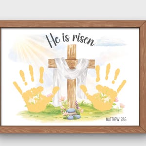 May include: A framed print featuring a wooden cross draped with white fabric, with the words "He is risen" above. Two yellow handprints flank the cross, set against a backdrop of a sunny sky and green grass. The text "Matthew 28:6" is at the bottom.