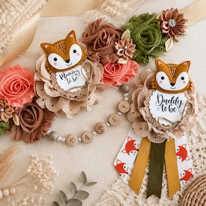 May include: A decorative sash and badge set featuring felt fox faces, fabric flowers in shades of coral, olive green, and brown, and wooden beads spelling KASEY. The fox faces are attached to scalloped lace circles with text reading Mommy to be and Daddy to be.