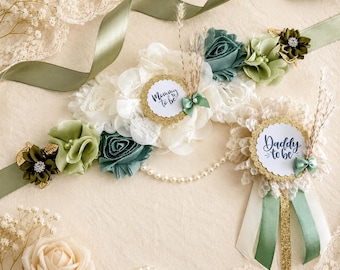 Peut inclure: Une ceinture de maternité et une broche assortie avec des fleurs en tissu blanc et vert sauge, des accents dorés pailletés et des perles. La ceinture porte l'inscription Mommy to be et la broche affiche Mommy to be en écriture bleue.