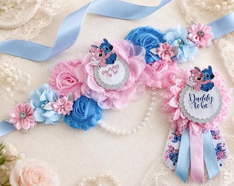 Op de afbeelding: Een babyshower sjerp en speld set met lichtblauwe en roze stoffen bloemen, kant en parelaccenten. De sjerp toont een rond label met de tekst Mommy to be en een Stitch en Angel afbeelding. De speld is voorzien van de tekst Daddy to be.