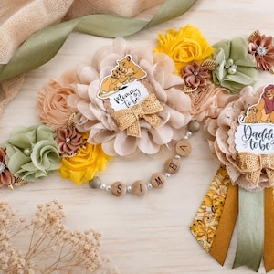 May include: A maternity sash and a badge featuring fabric flowers in beige, yellow, and sage green. The sash includes a cutout of a lioness and cub with the text Mommy to be, and the badge features a lion and cub with the text Daddy to be.