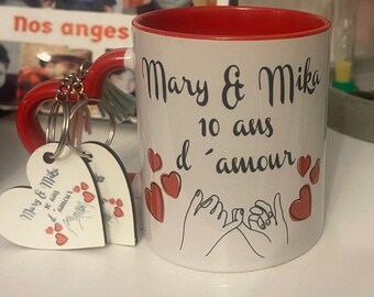 Può includere: Una tazza in ceramica bianca con un bordo e un manico rossi. La tazza ha un testo nero che recita "Mary & Mika 10 ans d'amour" ed è decorata con cuori rossi. La tazza è accompagnata da due portachiavi a forma di cuore con lo stesso testo e cuori rossi.