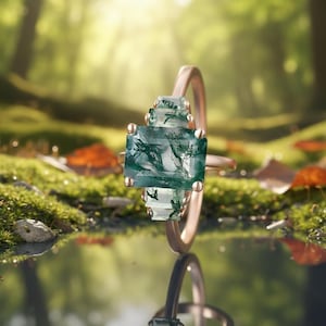 May include: A rose gold ring featuring a large, rectangular, green moss agate gemstone. The ring has a tiered design with smaller gemstones above the main stone. The ring is set against a blurred forest background, with a reflection in the foreground.