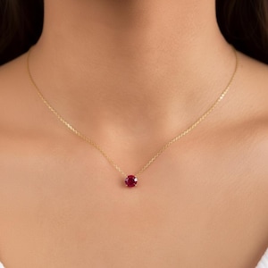 May include: A delicate gold necklace featuring a vibrant red gemstone pendant. The round-cut ruby is the focal point, suspended from a fine gold chain. The necklace is displayed against a skin tone background.