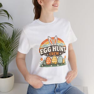 May include: White t-shirt with a colorful graphic design featuring the text "EGG HUNT CREW", bunny ears, a rainbow, and Easter eggs. The design is in a retro style, perfect for Easter celebrations.