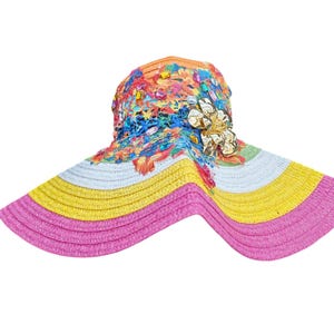 May include: A multi-coloured sun hat with a wide brim. The hat has a vibrant design with pink, yellow, white, and blue sections. The crown is decorated with colourful embellishments and a floral accent. This hat is designed to provide sun protection.