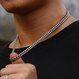 May include: A silver curb chain necklace. The necklace is made of interlocking links and is worn around the neck. The person in the image is wearing a black top and a silver ring.