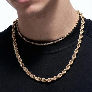 May include: Two gold-colored rope chain necklaces. The larger chain is thicker and hangs lower, while the smaller chain sits closer to the neck. The necklaces are worn over a black shirt.