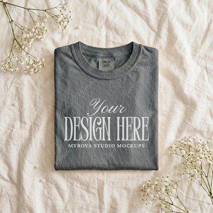 Puede incluir: Una camiseta gris con el texto "Your DESIGN HERE" en blanco, con "MYROVA STUDIO MOCKUPS" debajo. La camiseta está doblada sobre una tela color crema, con ramitas de flores blancas alrededor. La camiseta tiene una etiqueta Comfort Colors.