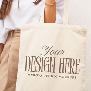 Puede incluir: Bolso tote de color crema con el texto marrón "Your DESIGN HERE" y "MYROVA STUDIO MOCKUPS" debajo. El bolso lo sostiene una persona que lleva una camiseta blanca y pantalones color beige. Se ve una pulsera dorada.