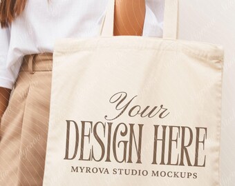 Puede incluir: Bolso tote de color crema con el texto marrón "Your DESIGN HERE" y "MYROVA STUDIO MOCKUPS" debajo. El bolso lo sostiene una persona que lleva una camiseta blanca y pantalones color beige. Se ve una pulsera dorada.