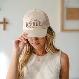 Puede incluir: Gorra de béisbol beige con el texto "Your Design Here" en letras marrones. La gorra es una maqueta de Myrova Studio Mockups. La gorra está siendo sostenida por una persona.