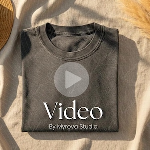 Puede incluir: Una camiseta gris oscuro doblada con cuello redondo. La palabra "Video" se muestra en blanco, junto con "By Myrova Studio". Un sombrero de paja y una planta seca están en el fondo.