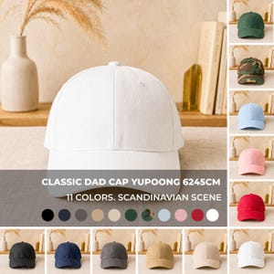 Puede incluir: Una gorra de béisbol blanca con una corona estructurada y visera curva. La gorra presenta una costura central, ojales para ventilación y un botón en la parte superior. El texto "CLASSIC DAD CAP YUPOONG 6245CM 11 COLORS. SCANDINAVIAN SCENE" está superpuesto en la imagen.