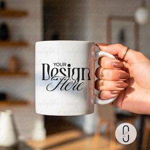 Puede incluir: Taza de cerámica blanca con el texto "YOUR Design Here" en escritura negra. La taza está sostenida por una mano con anillos dorados y esmalte de uñas marrón. La taza tiene un asa blanca.