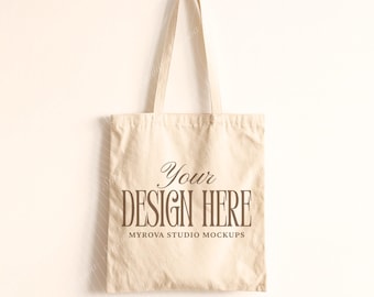 Puede incluir: Un bolso tote de lona beige con asas largas, colgado contra un fondo claro. La bolsa presenta el texto "Your DESIGN HERE" en una fuente elegante de color marrón oscuro, con "MYROVA STUDIO MOCKUPS" debajo.