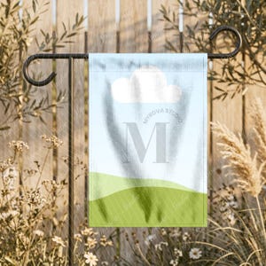 May include: A garden flag with a light blue sky background, white cloud, and green rolling hills. The flag has a large gray letter "M" and the text "MYROVA STUDIO". The flag is hanging on a black metal garden stake.