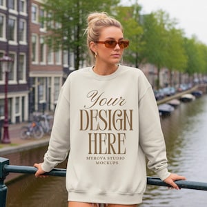 Puede incluir: Una sudadera crema con el texto "Your DESIGN HERE" en escritura marrón. La sudadera es una maqueta de Myrova Studio Mockups. La persona lleva gafas de sol y está de pie cerca de un canal.