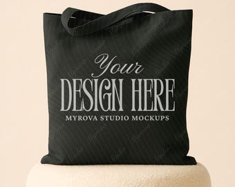 Puede incluir: Bolso tote negro con el texto "Your DESIGN HERE" en fuente serif. El bolso tiene un patrón de rayas y un asa. Las palabras "MYROVA STUDIO MOCKUPS" están debajo.