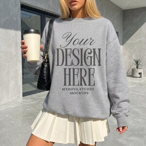 Puede incluir: Una sudadera gris con el texto "Your Design Here" se combina con una falda plisada blanca. También se ven un bolso negro y una taza de café. La sudadera es una maqueta de Myrova Studio Mockups.