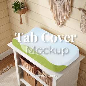 May include: A baby changing mat cover with a blue and green design, displayed on a white changing table. The text "Tab Cover Mockup" is visible on the cover. Wicker baskets and decorative items are in the background.