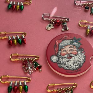 May include: A collection of gold-tone safety pins featuring various Christmas-themed charms including miniature light bulbs, mittens, stockings, bells, and snowflakes, alongside a round red coaster with a Santa Claus illustration.
