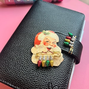 Op de afbeelding: Een zwart getextureerd notitieboek met een uitgeknipte kerstman bevestigd met een gouden veiligheidsspeld met vier glitterende rode, gele en groene kerstlampjes, plus een bijpassende kerstlampjesclip aan de zijkant.