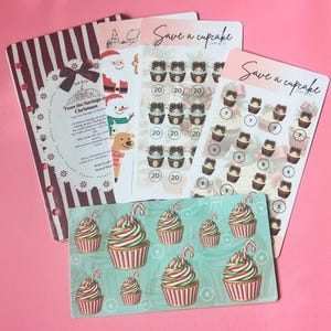 May include: A collection of four paper savings challenge trackers. One features a maroon striped design with text, another shows Christmas characters, and two are titled Save a cupcake with cupcake illustrations and numbered circles.