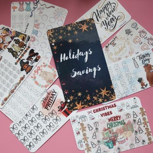 May include: A collection of decorative planner inserts with various designs and text. Includes inserts with text like "Holidays Savings", "Happy New Year", "Black Friday", and "Merry Christmas". The inserts are on a pink surface.