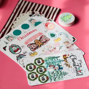Op de afbeelding: Een set kerstthema spaarchallenge-kaarten met illustraties van peperkoek, sneeuwpoppen en rendieren. De kaarten bevatten cirkelvormige vakjes met bedragen zoals $5 en $10, en de tekst Merry Christmas.