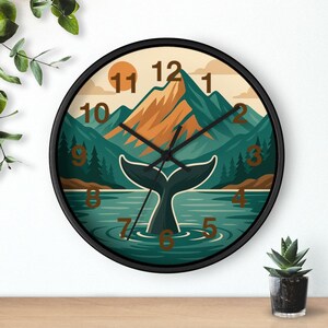 May include: A round wall clock with a black frame. The clock face features a landscape design with a whale tail in the water, mountains, and a setting sun. The numbers are in a brown font.