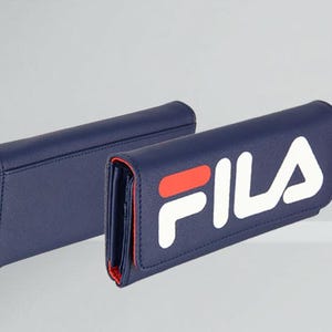 Pu&ograve; includere: Due portafogli blu navy con finiture rosse. Un portafoglio presenta il logo "FILA" bianco e rosso. I portafogli sono esposti su una superficie bianca su sfondo grigio chiaro. I portafogli sembrano essere in pelle.