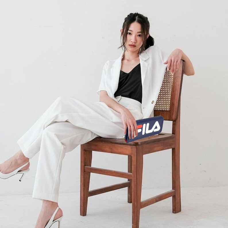 Pu&ograve; includere: Una persona con una giacca bianca, un top nero e pantaloni bianchi, seduta su una sedia di legno. Una pochette blu navy con il logo bianco "FILA" &egrave; tenuta in grembo. Lo sfondo &egrave; un muro bianco.