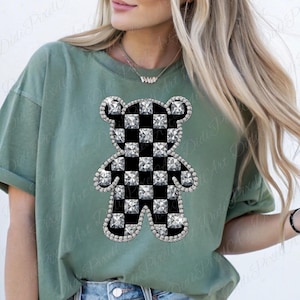 May include: Sage green t-shirt featuring a teddy bear design. The bear has a black and white checkered pattern and is outlined with sparkling rhinestones. The shirt has short sleeves and a relaxed fit.