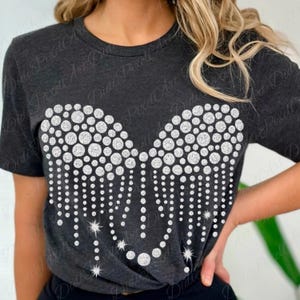 May include: Dark gray t-shirt featuring a sparkling, heart-shaped design made of white rhinestones. The design includes various sizes of circular rhinestones and dangling strands, with star accents. The shirt is a crew neck style.