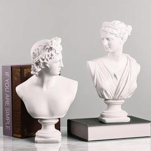 May include: Two white plaster busts of classical figures. One bust has a laurel wreath, the other has a draped garment. The busts are on pedestals, and one is on a book. A book with the text "IF YOU ARE SIMPLE" is also visible.