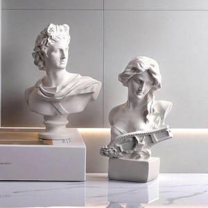 Può includere: Due busti in gesso bianco di figure classiche. Uno è un busto maschile con capelli ricci e un drappeggio. L'altro è un busto femminile che tiene una lira. Entrambi sono su piedistalli bianchi, su sfondo neutro.