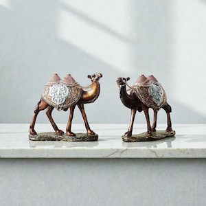 Può includere: Due statuette decorative di cammelli. I cammelli sono marroni con selle ornate nei toni dell'oro, dell'argento e del rosa. Si trovano su una base rettangolare. Lo sfondo è una parete grigio chiaro e una superficie bianca.
