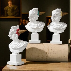 Può includere: Tre sculture di busti bianchi con accenti di coni gelato. I busti sono su piedistalli bianchi, con un cono che sembra sciogliersi. I coni hanno vari colori, tra cui marrone, rosso e blu. I busti sono esposti su una superficie di legno.