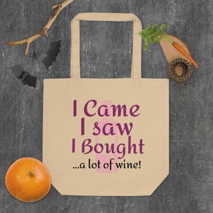 Puede incluir: Bolso tote beige con el texto morado "I Came I Saw I Bought... a lot of wine!". Un gráfico de botella de vino rosa está entre "Saw" y "Bought". La bolsa está sobre una superficie gris con una calabaza, murciélagos decorativos y una zanahoria.