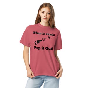 Puede incluir: Camiseta de color rosa polvoriento con el texto negro "When in Doubt Pop it Out!". El gráfico muestra una botella de champán descorchándose con un sacacorchos. La camiseta es de estilo casual y cómodo.