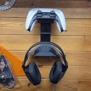 May include: A black wall-mounted storage rack holding a white and black game controller at the top and a pair of black over-ear gaming headphones on the lower support hook. The rack features a vertical design with two distinct cradles for gaming gear.