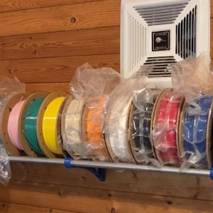Puede incluir: Una fila de bobinas circulares de filamento en varios colores, incluyendo amarillo, morado, rosa, turquesa, naranja, blanco, negro, rojo, azul y marrón, cada una envuelta individualmente en plástico transparente sobre un estante metálico.