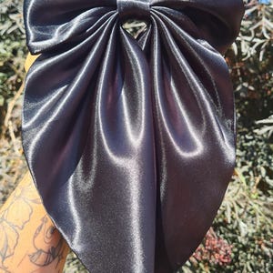 May include: A large, dark charcoal grey hair bow crafted from a smooth, lustrous satin fabric. The accessory features a wide, gathered top section with a central knot and a long, pointed, draped tail that hangs downwards in a soft, flowing shape.