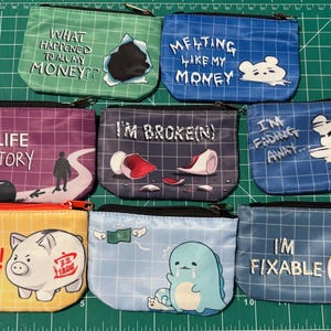 May include: A collection of small, zippered pouches with various designs and text. The pouches feature phrases like "What happened to all my money?" and "Give me money!" along with cartoon illustrations and a grid pattern background.