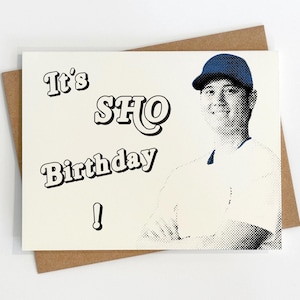 May include: A birthday card with the text "It's SHO Birthday!" in a retro font. The card features a halftone image of a person wearing a baseball cap and a white shirt. The card is on a brown envelope.