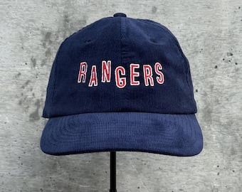 May include: Navy blue corduroy baseball cap with the word "RANGERS" embroidered in red and white. The cap has a curved brim and a rounded crown. A classic style, suitable for everyday wear.