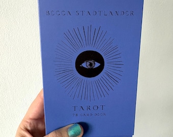 Może przedstawiać: Prostokątne niebieskie pudełko na karty tarota z napisem BECCA STADTLANDER na górze i TAROT 78 CARD DECK na dole. Na środku pudełka znajduje się czarna okrągła grafika oka otoczonego promieniującymi cienkimi czarnymi liniami.
