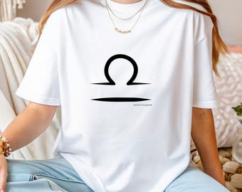 Puede incluir: Camiseta blanca con un símbolo negro del signo zodiacal Libra. El diseño presenta un símbolo de balanza estilizado sobre una línea horizontal. La camiseta se combina con vaqueros azul claro. El texto "collectivespirit" está debajo.