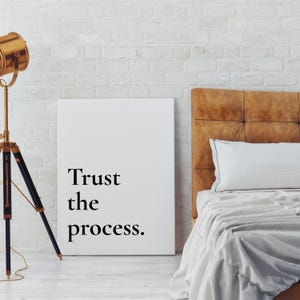以下が含まれることがあります： 「Trust the process」の黒い文字が書かれた白いキャンバスプリント。白いレンガの壁に立てかけられており、茶色のヘッドボード、白い枕と毛布のあるベッドの近くにあります。左側にはヴィンテージスタイルのランプがあります。